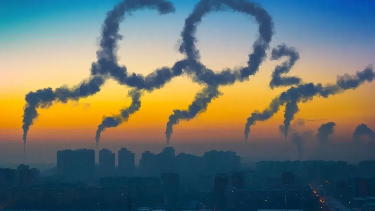 Dym z kominów układa się w napis "CO2", symbolizując globalną emisję dwutlenku węgla nad miastem o zachodzie słońca.