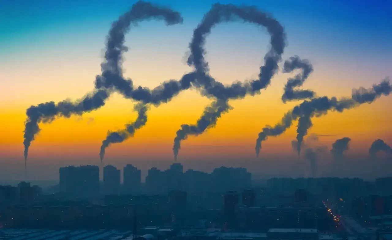 Dym z kominów układa się w napis "CO2", symbolizując globalną emisję dwutlenku węgla nad miastem o zachodzie słońca.