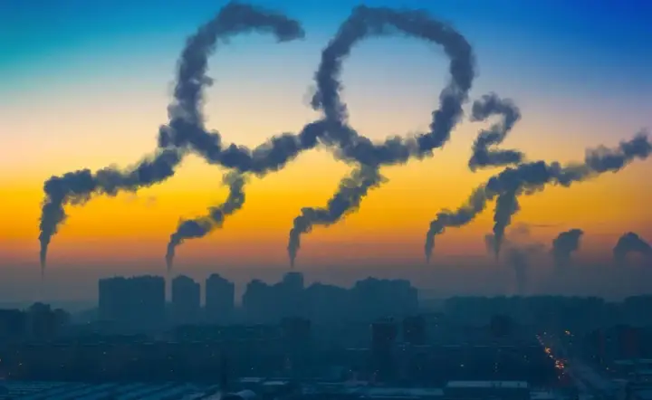 Dym z kominów układa się w napis "CO2", symbolizując globalną emisję dwutlenku węgla nad miastem o zachodzie słońca.